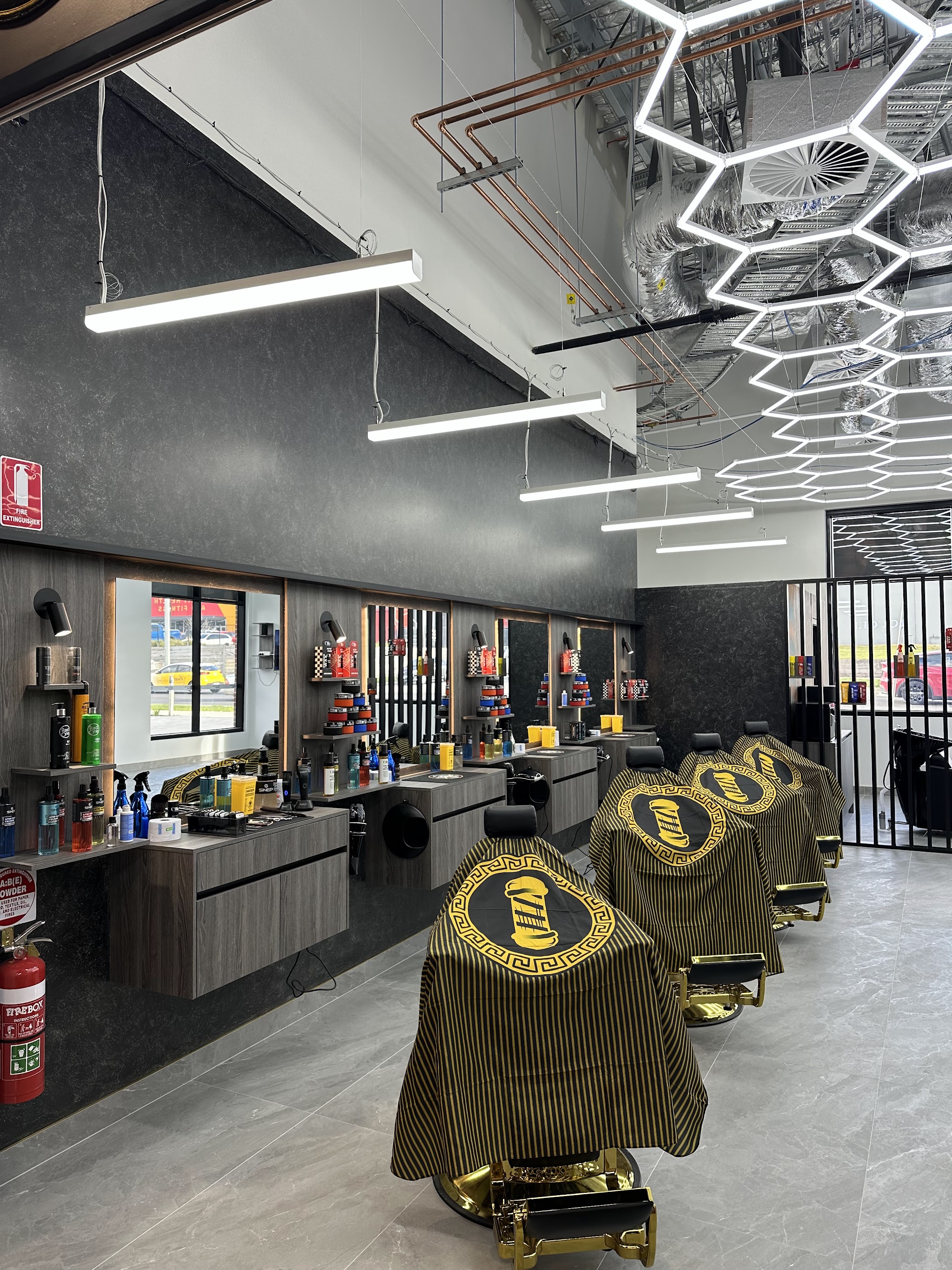 Inside the shop: gold barber chairs in black and gold capes lined up beside backlit mirrors under a hexagon light grid